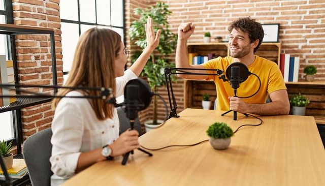High-fiving podcast hosts recording together in bright brick studio with table microphones