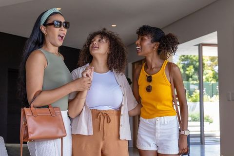 Diverse female friends greeting and laughing in modern home foyer with sunglasses