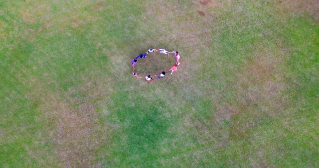 Children forming a circle by holding hands outdoors on green grass. Perfect for concepts relating to teamwork, unity, friendship, and outdoor activities. Could be used in promotional materials for parks, schools, or community events.