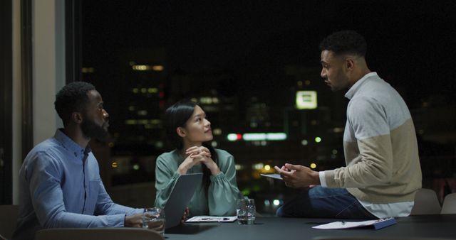 Three diverse business professionals having a discussion late at night in a modern office with a night cityscape background. One person is standing while two are seated, creating a collaborative atmosphere. Ideal for concepts of teamwork, business meetings, brainstorming sessions, hard work, and dedication.