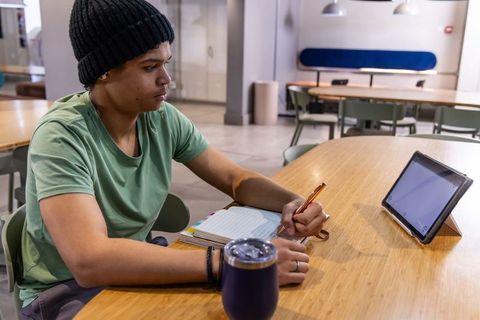 African american teen studying and writing at campus lounge with tablet and tumbler