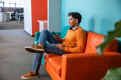 Asian man relaxing on orange couch in modern office lounge using smartphone and headphones