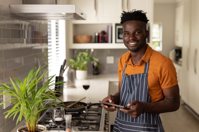 Portrait of smiling african american young man using digital tablet while cooking in kitchen at home. cooking, unaltered, lifestyle, home and wireless technology concept.