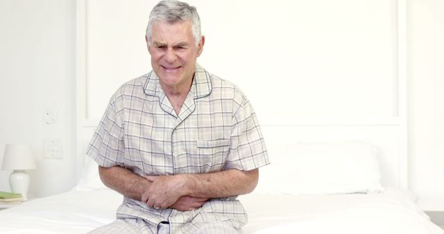 Elderly man sitting on bed clutching his stomach in pain. Useful for illustrating medical conditions, health issues, senior care, or home healthcare scenarios. Can be used in health-related content, articles on common illnesses among seniors, or advertisements for medical and healthcare services.