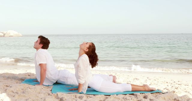 A young Caucasian man and woman practice yoga on a serene beach, with copy space. Their pose promotes relaxation and wellness against the tranquil backdrop of the ocean.