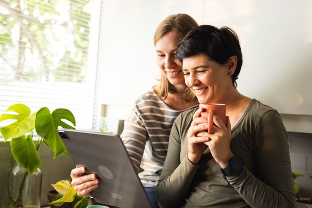Smiling caucasian lesbian couple watching video over digital pc while having coffee in kitchen. Drink, wireless technology, unaltered, love, togetherness, homosexual, lifestyle and home concept.