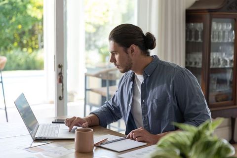 Man working from home at sunlit wooden table using laptop taking notes with coffee mug