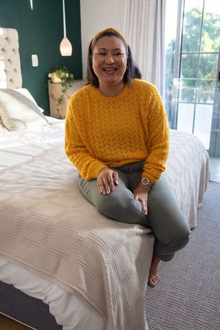 Smiling woman sitting on bed in cozy sunlit bedroom wearing mustard sweater and olive pants