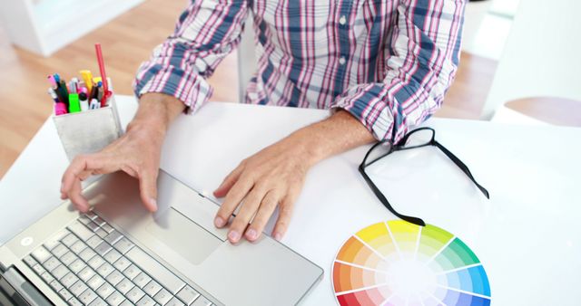 A middle-aged Caucasian man works on a graphic design project using a laptop, with copy space. His workspace includes a color wheel, suggesting a focus on creativity and design aesthetics.