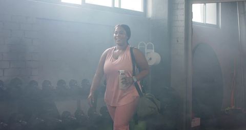 Mid-adult african american woman walking in gym wearing pink activewear carrying duffel and shaker