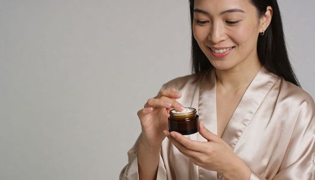 Smiling asian woman applying moisturizer from amber jar wearing satin robe for skincare