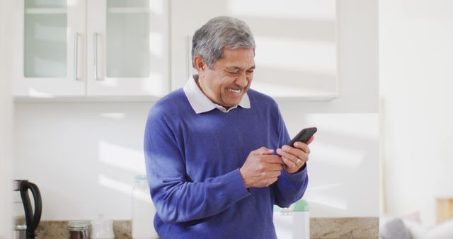 Image of happy senior biracial man using smartphone in kitchen, smiling and laughing. Retirement, communication, inclusivity and senior lifestyle concept.
