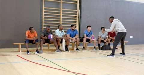 Coach coaching diverse male volleyball team on bench in indoor gym during training