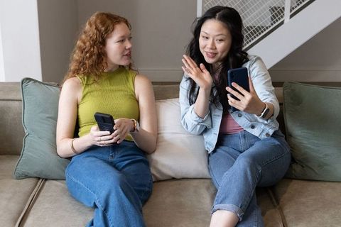 Two friends sitting on sofa waving and texting on smartphones casual home conversation