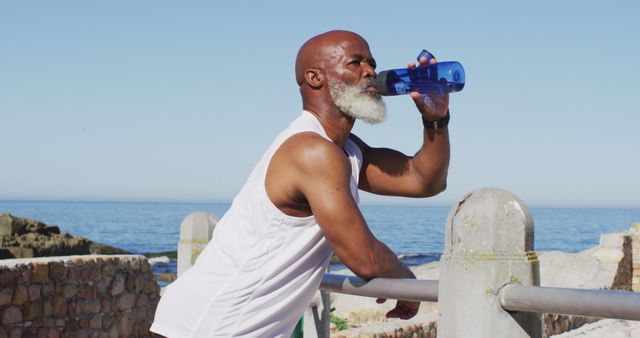 Senior man in white tank top drinking water from a blue bottle and resting during outdoor workout by the beach. Ideal for content promoting hydration, fitness routines, or a healthy, active lifestyle for older adults. Suitable for health and wellness articles, exercise blogs, and advertisements emphasizing outdoor activities and beachside workouts.