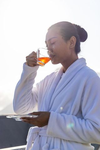 Sunlit morning ritual: asian woman sipping tea in plush white robe on balcony