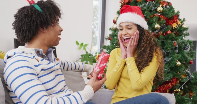 Image of happy diverse couple exchanging christmas gift and embracing at home. Christmas, celebration, happiness, tradition and inclusivity concept.
