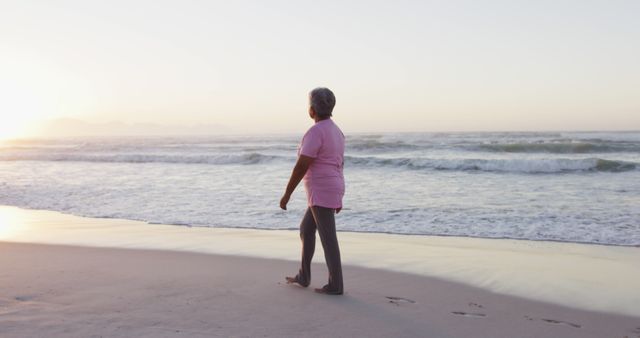 Elderly woman walking by the ocean as the sun sets, wearing casual clothing. Ideal for themes of relaxation, retirement, enjoying life, mindfulness, and health. Can be used in travel brochures, lifestyle blogs, wellness articles, and marketing materials promoting serene locations.