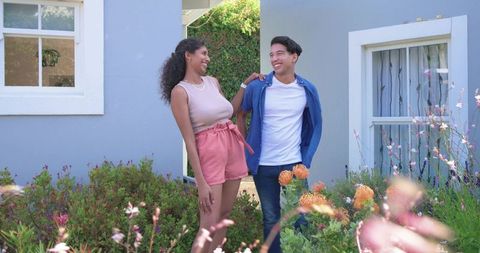 African american couple smiling in sunlit garden by blue house wearing casual clothes