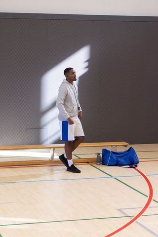 African american coach standing courtside holding stopwatch with blue duffel bag in gym