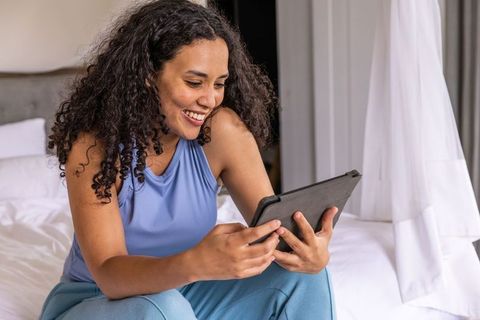 African american woman smiling and holding tablet on bed, cozy relaxed morning routine