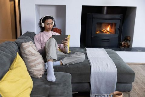 Nonbinary person relaxing on gray sofa with tablet and headphones by cozy fireplace
