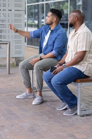 African american men sitting on urban bench pointing while checking smartphone and talking
