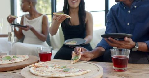 Friends sharing pizza in sunlit living room passing slices and toasting with drinks