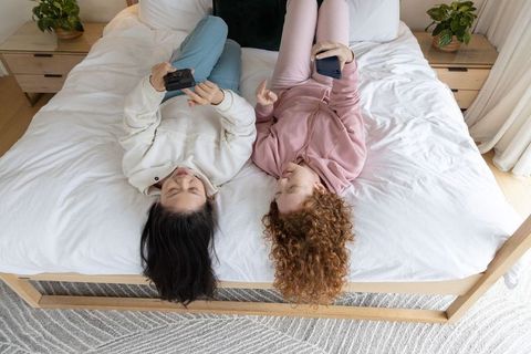 Two friends lying on bed using smartphones laughing overhead lifestyle portrait