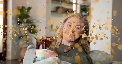 Woman smiling while sipping coffee on cozy sofa with warm golden bokeh