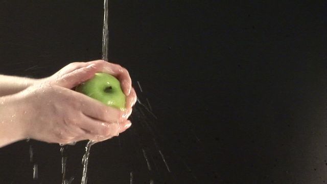 Hands are shown cleaning a green apple under running water against a dark background. Ideal usage for food safety, healthy lifestyle, kitchen hygiene, or clean eating content. Can be used in blogs, health-related articles, food preparation guidelines, and advertisements promoting fresh produce and cleanliness.