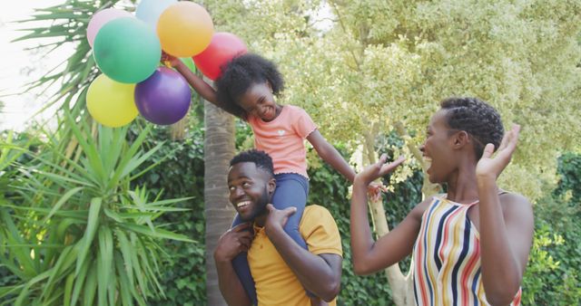 Happy african american parents and daughter holding balloons in garden. Lifestyle, domestic life, family, and togetherness.