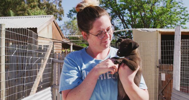This image depicts a woman in glasses holding a small puppy outdoors in an animal shelter setting. She is bonding with the puppy, indicating a sense of care and compassion, ideal for use in content related to pet adoption, animal rescue awareness, and promoting animal shelters. The natural lighting and fence backdrop highlight an authentic shelter environment, perfect for campaigns and advertisements centered around pet adoption.
