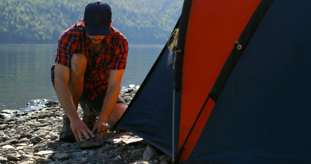 Man enjoying outdoor activity by setting up tent on rocky shore near a peaceful lake with mountains in the background. Perfect for travel, adventure, and nature-related content.