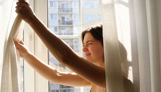 Sunlit young woman opening apartment curtains at morning window with city high-rise view