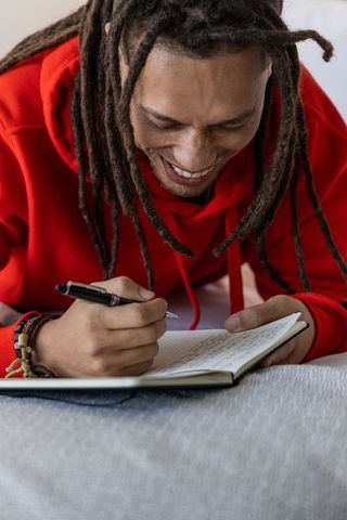 African american man writing in journal smiling on bed wearing red hoodie lifestyle