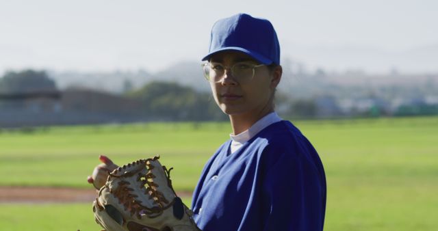 This image features a young female baseball player in a blue uniform and cap, standing on a baseball field under bright sunlight. She is holding a baseball glove, looking towards the camera with a serious expression. The well-lit outdoor setting and athletic context make this image suitable for sports-related articles, promoting women's participation in sports, athletic gear advertisements, or youth baseball programs.