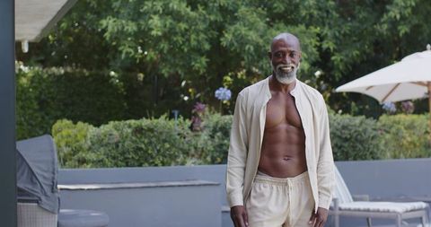 Fit senior man standing and smiling on patio wearing unbuttoned beige shirt and shorts