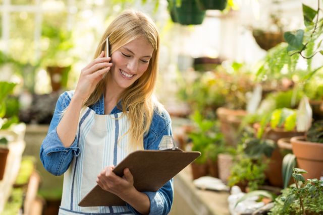 Young female gardener talking on cellphone while looking at clipboard in greenhouse