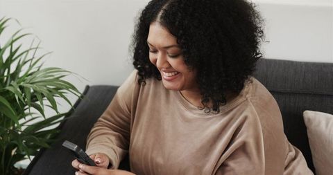 Smiling African American woman checking smartphone on cozy sofa, relaxed home lifestyle