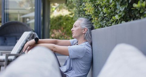 Senior woman relaxing on outdoor patio bench wearing smartwatch beside lush garden hedge