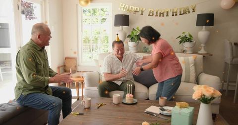 Mature man celebrating birthday at home with small cake, family clapping and gifts
