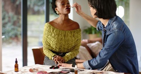 Makeup artist applying lip color with brush to black woman client in cozy sunlit room