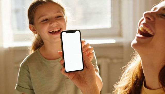 Mother laughing with daughter while holding smartphone showing blank white screen in sunlight