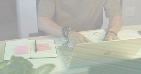 Man typing on laptop with notebook and pink sticky notes while checking pie chart on desk