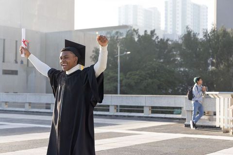 Joyful graduate celebrating on urban campus holding diploma wearing mortarboard and gown