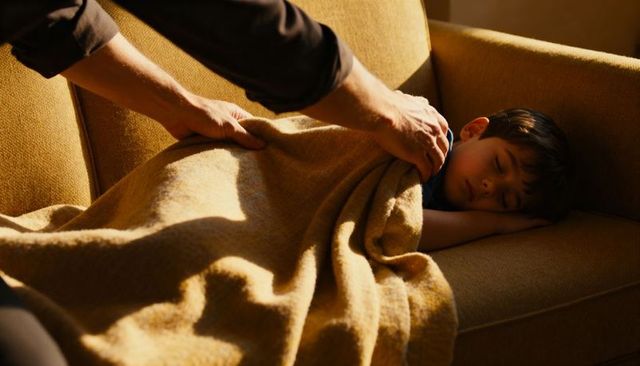 Parent tucking sleeping boy under wool blanket on sunlit sofa during cozy late afternoon