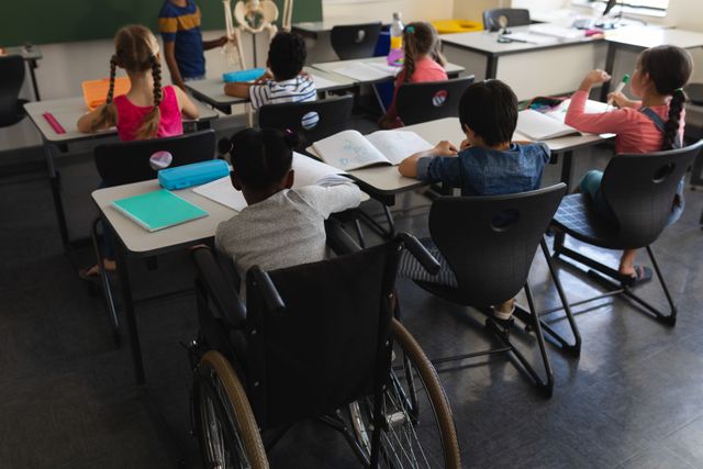 Rear view of disable schoolboy with classmate studying and sitting at desk in classroom of elementary school