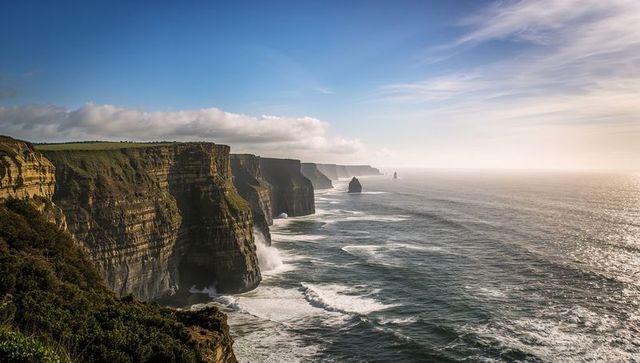 Dramatic layered coastal cliffs rising over churning ocean with sea stacks at sunrise
