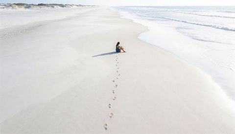 Sitting woman in dark tank top leaving footprints on expansive pale sand shoreline